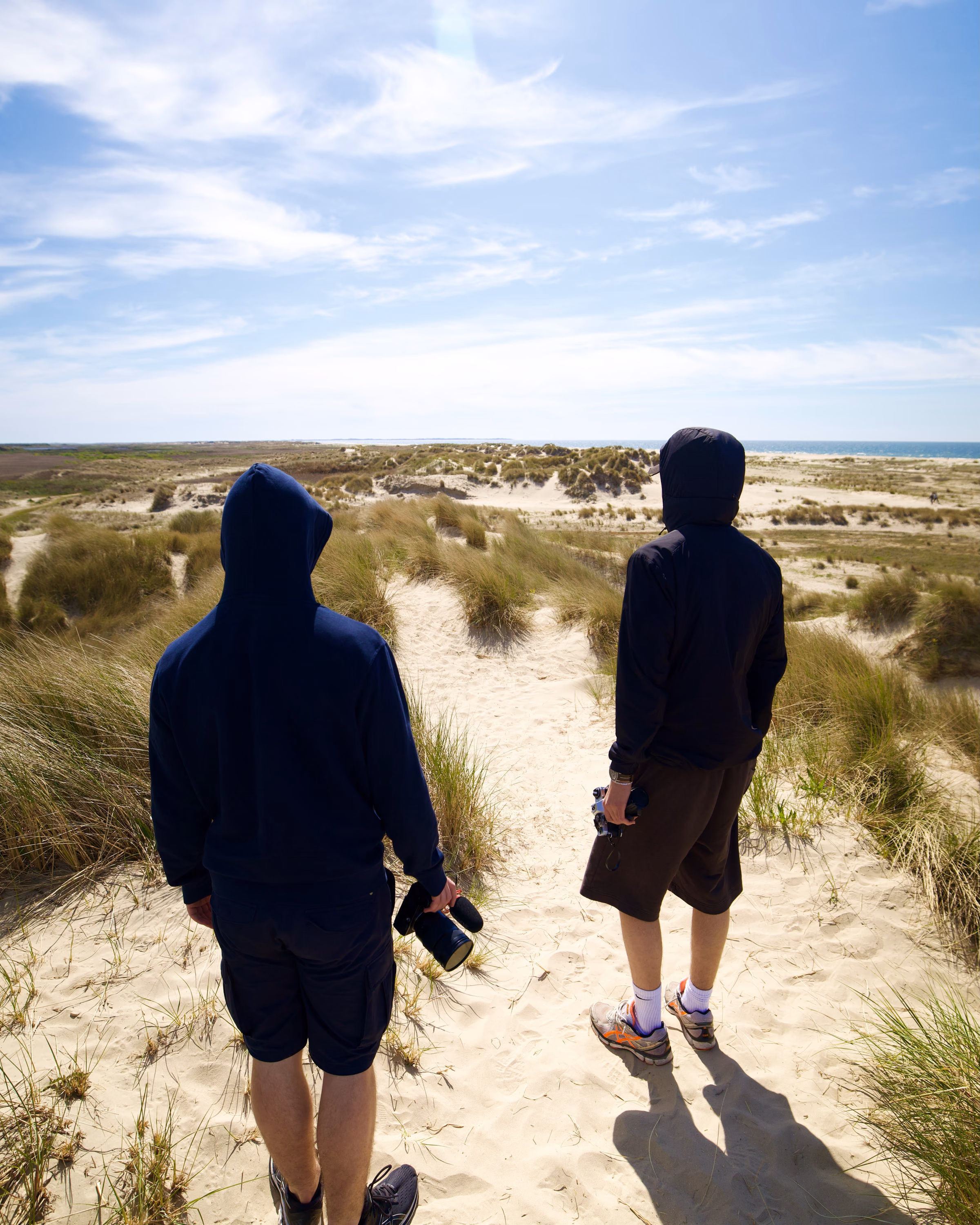Two filmmakers carrying cameras across coastal sand dunes